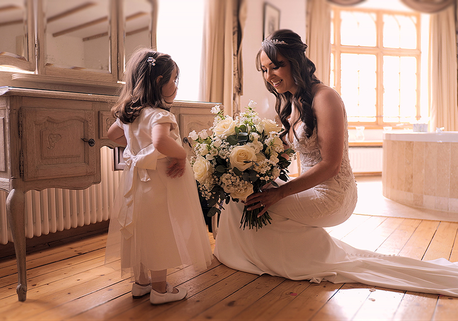 Bride crouches beside flower girl holding large bouquet of cream flowers